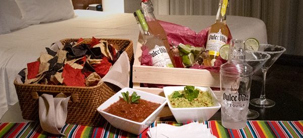Table with basket of various chips, salsa, bottled tequila, and martini glasses with a salt rim